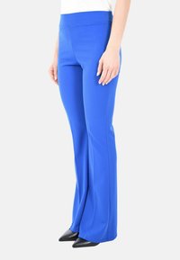 Donna che indossa pantaloni a campana aderenti di un vivace blu brillante e scarpe nere con punta affusolata, in piedi davanti a uno sfondo bianco semplice.