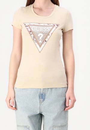 Kobieta w beżowej koszulce z krótkim rękawem z dużym trójkątnym logo Guess oraz jasnoniebieskich dżinsach.