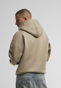 Beige oversized hoodie met een trekkoordkap, wijde mouwen en geribbelde manchetten, gecombineerd met lichtblauwe denim jeans met decoratieve stiksels.