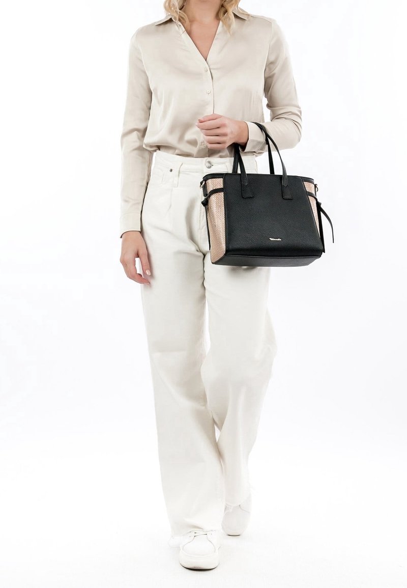 Femme portant un chemisier crème et un pantalon, tenant un sac fourre-tout noir et beige, marchant vers l'avant sur fond blanc.