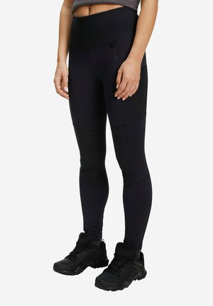 Frau trägt schwarze Leggings mit hoher Taille und Seitentaschen sowie schwarze Wanderschuhe, steht vor weißem Hintergrund.