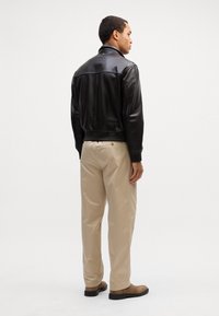 Giubbotto di pelle nera con un design aderente, polsini e colletto a coste, abbinato a pantaloni beige e stivaletti alla caviglia marroni. Sfondo neutro.