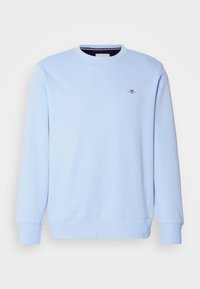 Sweat-shirt bleu clair à manches longues avec col rond, poignets et ourlet côtelés, comportant un petit blason brodé sur la poitrine gauche.