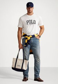 Hvit bomulls-T-skjorte med «POLO»-logo, marineblå og gulstripet genser bundet rundt midjen, blå jeans og beige handlenett med marineblå detaljer.