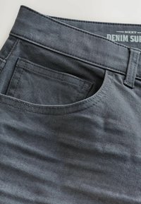 Tuvumā redzama tumši pelēka džinsu auduma bikses, kuras rāda priekšējo kabatu, jostas cilpu un daļu no jostasvietas ar tekstu "DENIM SURF" iekšpusē.