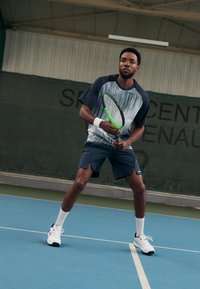 Tennisspieler bereit mit einem grünen Schläger, trägt ein blau-weiß gestreiftes Shirt, dunkle Shorts und weiße Turnschuhe auf einem blauen Platz.
