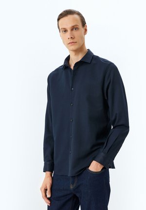 Marineblå skjorte med knaplukning og krave, lange ærmer og en glat tekstur, parret med mørk denim jeans. Minimalistisk design uden synlige mønstre.