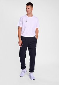 Camiseta blanca de manga corta con un logo negro y pantalones con cintura elástica negra. El modelo lleva zapatillas blancas. Diseño simple y limpio.