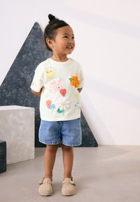 Light yellow short-sleeve t-shirt with colorful embroidered designs including flowers and cartoon characters, paired with denim shorts and beige shoes.