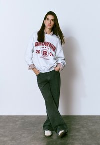 Sudadera gris con "BROWNIE 2008 COLLEGE CREW" en burdeos, combinado con una camisa de rayas. Pantalones de pierna ancha verde oscuro y zapatillas blancas.