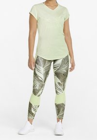 Ljust grön kortärmad t-shirt med v-ringning och en diskret logotyp, tillsammans med åtsittande leggings med kamouflagemönster och vita bladmönster.