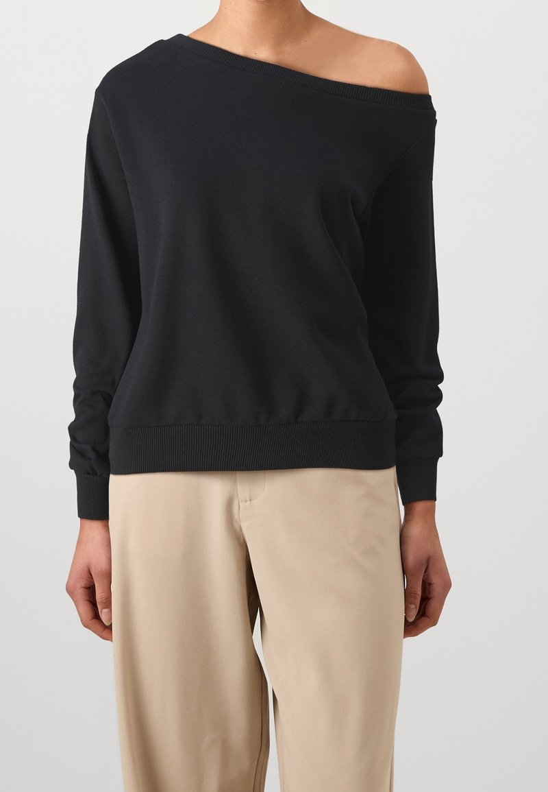Femme portant un top noir à épaules dénudées et manches longues, associé à un pantalon beige ample sur un fond uni.