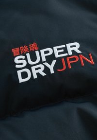 Čierna tkanina s vyrazeným bielym a červeným textom. Dizajn obsahuje názov značky "SUPER DRY" s japonskými znakmi nad ním v červenej farbe.
