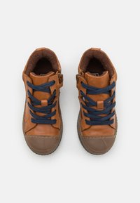 Baskets en cuir marron avec des lacets bleu marine, semelles en caoutchouc marron et un intérieur texturé et rembourré. Fermeture éclair sur le côté pour un accès facile.