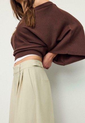 Femme ajustant un pantalon plissé beige à taille haute, portant un pull marron à épaules dénudées et un sous-pull blanc, sur un fond uni.