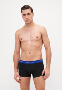 Fekete boxert nadrág kék derékrésszel, amelyen a "TOMMY HILFIGER" logó található. Simító anyag és testhezálló dizájn. A modell a karját a test mellett tartja.