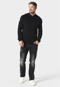 Sort genser med glidelås i halsen og lange ermer, kombinert med falmede svarte jeans og hvite joggesko. En enkel, moderne design med myk tekstur.