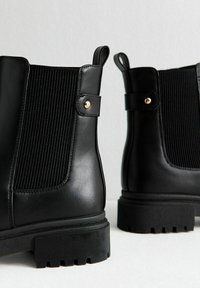 Botas negras de cuero hasta el tobillo que presentan paneles acanalados, detalles en dorado y una suela gruesa texturizada para mejorar el agarre.