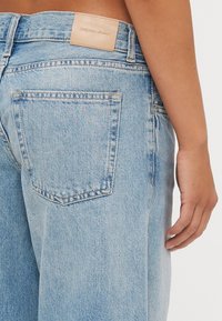 Calças de ganga de denim azul claro com uma cintura alta, apresentando um rótulo de couro na parte de trás, cinco bolsos e uma textura ligeiramente desgastada.
