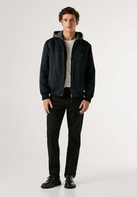 Chaqueta bomber negra con forro de sudadera gris, cremallera frontal y puños acanalados, combinada con pantalones negros y zapatos negros, sobre un fondo neutro.