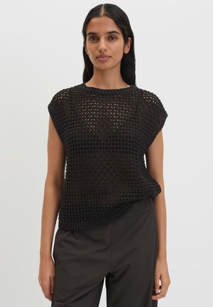 Femme portant un haut noir en crochet sans manches sur un pantalon noir, debout devant un fond gris clair uni.