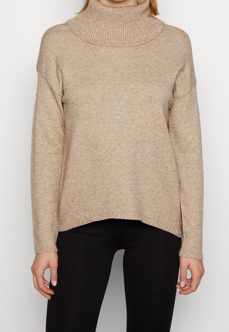 Femme portant un pull à col roulé beige avec col et poignets côtelés, associé à un pantalon noir, visible du cou aux hanches.