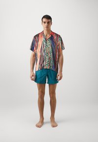 Kortærmet, flerfarvet skjorte med et livligt abstrakt mønster, parret med ensfarvede teal shorts. Modellen står barfodet mod en ensfarvet baggrund.
