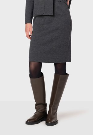 Bottes en cuir marron jusqu'au genou avec une semelle plate, portées avec une jupe ajustée grise foncé et des collants noirs. Le matériau a une texture lisse.