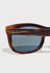 Ochelari de soare din tortoiseshell maro cu lentile albastre. Prezintă un design îndrăzneț și unghiular, cu marca Ray-Ban pe lateral. Finisaj lucios.