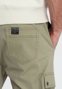 Męska ręka w kieszeni beżowych spodni cargo, z widoczną etykietą tylnej kieszeni i fragmentem białej podkoszulki z prążkami.