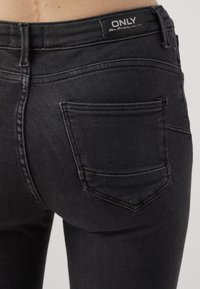 Jeans de mezclilla negra con un diseño ajustado, que cuentan con detalles en los bolsillos traseros y una etiqueta de cintura visible. Tejido texturizado con un ligero brillo.