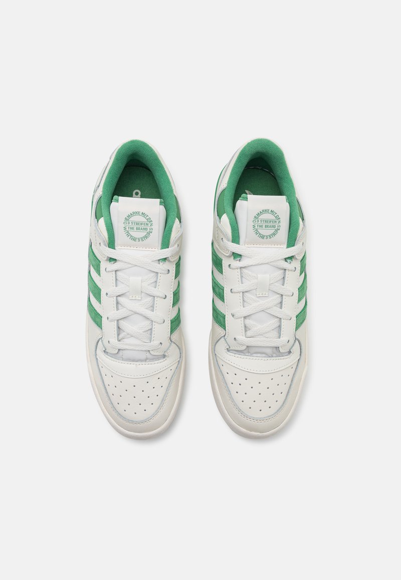 Zapatillas blancas con rayas verdes, área del dedo del pie perforada, cuello acolchado y cordones blancos. Logo circular en la lengüeta. Suela de goma plana.