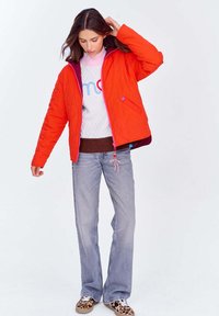 Kobieta poprawiająca ciemnobrązowe włosy, ubrana w jasnopomarańczową pikowaną kurtkę, białą bluzę, jasnoniebieskie dżinsy i trampki w leopardzi wzór, stoi na tle białym.