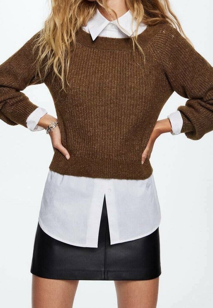 Femme portant un pull en maille marron sur une chemise blanche avec les poignets apparents, associée à une minijupe en cuir noire, mains sur les hanches.