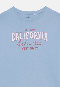 Šviesiai mėlynas medvilninis marškinėlis su apvaliu kaklu. Papuoštas raudonu ir baltu spaudiniu: „CALIFORNIA EST. 1982 Golden State WEST COAST.“
