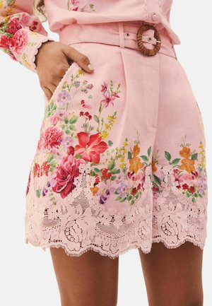 Hand in pocket, wearing pink shorts with multicolored floral print and lace trim, featuring a brown circular belt buckle.