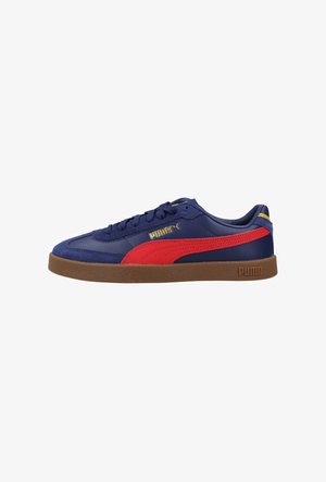Zapatilla Puma azul marino con franja roja, suela de goma marrón, logo dorado de Puma en el lateral y diseño de corte bajo.