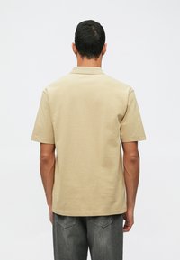 Polo beige en coton à manches courtes, avec un col et un ourlet droit. Porté avec un pantalon gris foncé, vu de l'arrière.