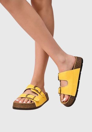 Jambes croisées, portant des sandales jaunes avec deux brides à boucle et des semelles en liège sur un fond clair uni.