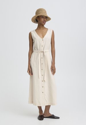 Donna che indossa un vestito midi beige senza maniche con bottoni sul davanti e vita annodata, un cappello di paglia intrecciata e scarpe scure senza lacci, in piedi su uno sfondo neutro.