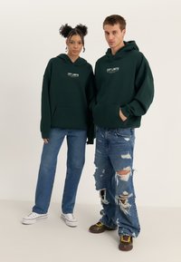 Green hooded sweatshirts with "OFF LIMITS" text, paired with distressed blue jeans. Model wears white sneakers; second model wears brown sneakers.