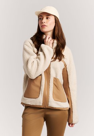 Young woman wearing a beige fleece jacket with brown pockets and panels, brown pants, and a light beige cap, looking to the side.