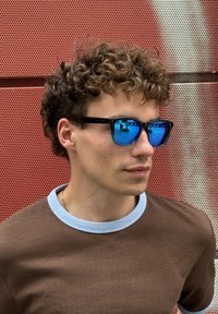 Ochelari de soare rectangulari negri cu lentile reflective albastre, purtați de o persoană în cămașă maronie cu guler albastru deschis. Fundal texturat.