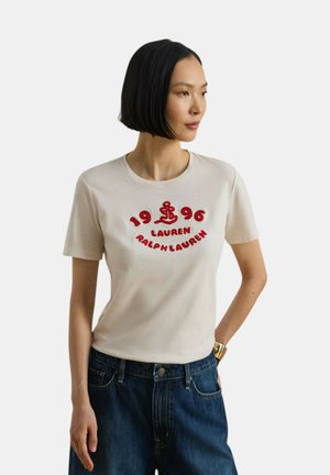 Donna con capelli neri lisci taglio bob indossa una maglietta crema a maniche corte con scritta rossa "1996 Lauren Ralph Lauren" e un'ancora, abbinata a jeans blu.