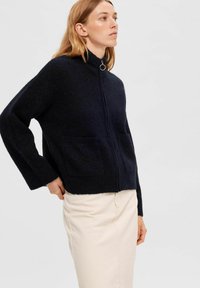 Marineblauwe zip-up trui met een hoge kraag, twee voorkennis en een zachte, gestructureerde stof; gecombineerd met een crèmekleurige rok.
