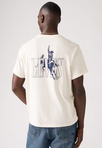 Krems krāsas T-krekls ar lielu tumši zilu grafiku, attēlojot kovboju uz zirdziņa, un zem tā ir teksts "LEVI'S". Īsas piedurknes, standarta piegriezums.