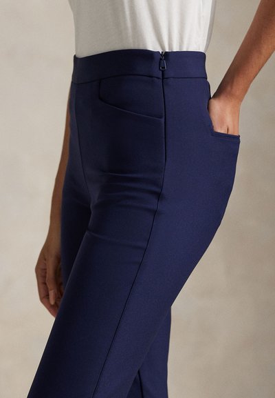 Persoon draagt aangesloten marineblauwe broek met zijzakken en een rits in de taille, zichtbaar van de taille tot halverwege het dijbeen.