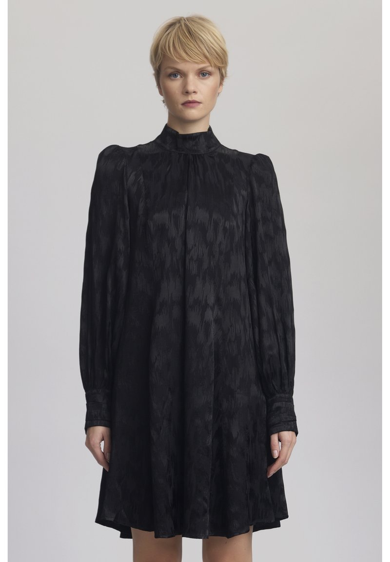Schwarzes Kleid mit langen, gepufften Ärmeln und hohem Kragen. Hergestellt aus strukturiertem Stoff mit subtilen Mustern. A-Linien-Form, endet oberhalb des Knies.