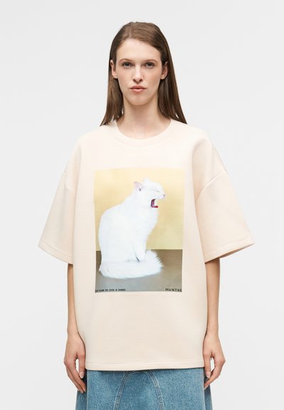 Μπεζ υπερμεγέθη μπλουζάκι από απαλό ύφασμα, με μπροστινό τυπώμα μιας λευκής γάτας που χασμουριέται πάνω σε χρυσό φόντο.