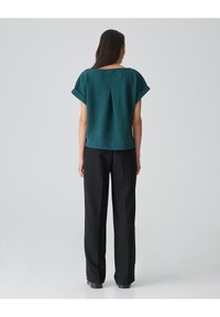 Teal gestructureerde top met een brede halslijn en korte mouwen, gecombineerd met zwarte geklede broeken. De top heeft een middenplooi detail.